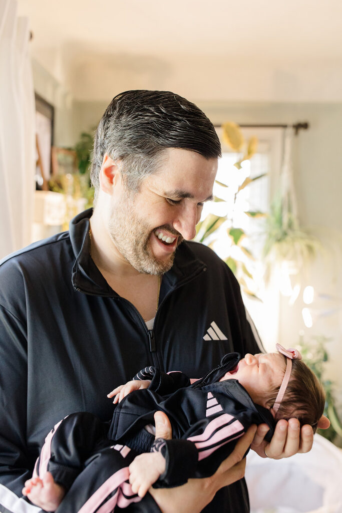 Father holds his newborn in a matching black and pink tracksuit during an in-home session, showing that personal style can be part of what to wear for authentic newborn photos by a Cleveland family photographer.