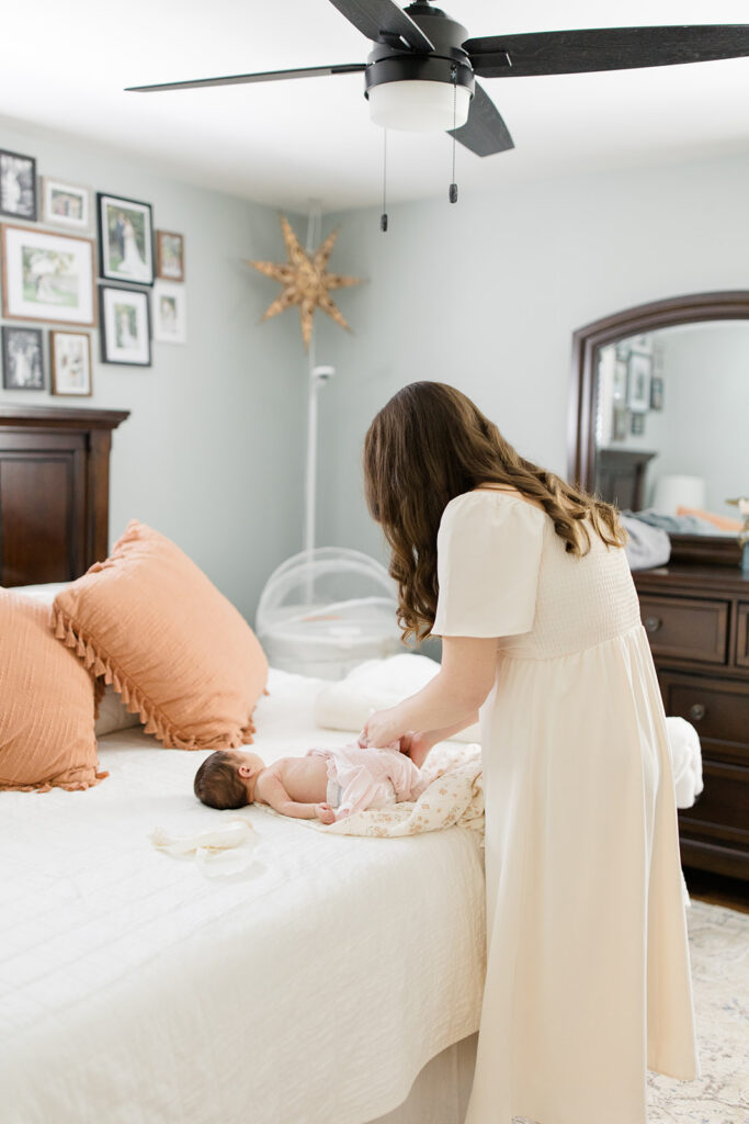 Mother dresses her newborn on a bed in a soft neutral dress, providing timeless what to wear inspiration for cozy in-home newborn photos by a Cleveland family photographer.