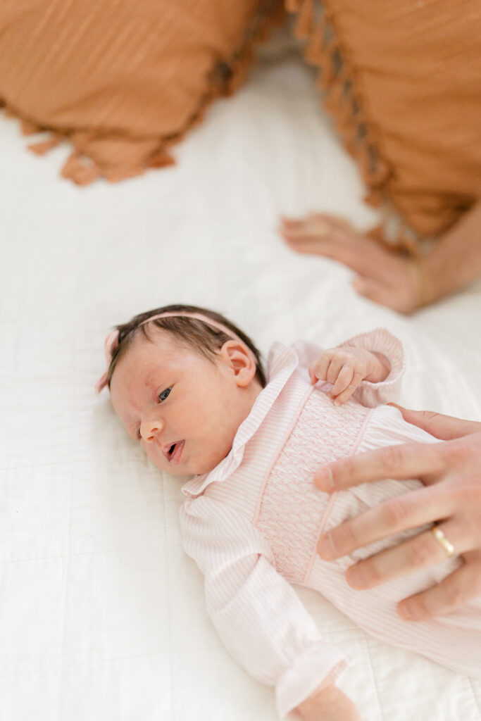 Newborn in a pink smocked outfit and bow headband lying on a bed, highlighting classic what to wear ideas for natural, in-home newborn photos.