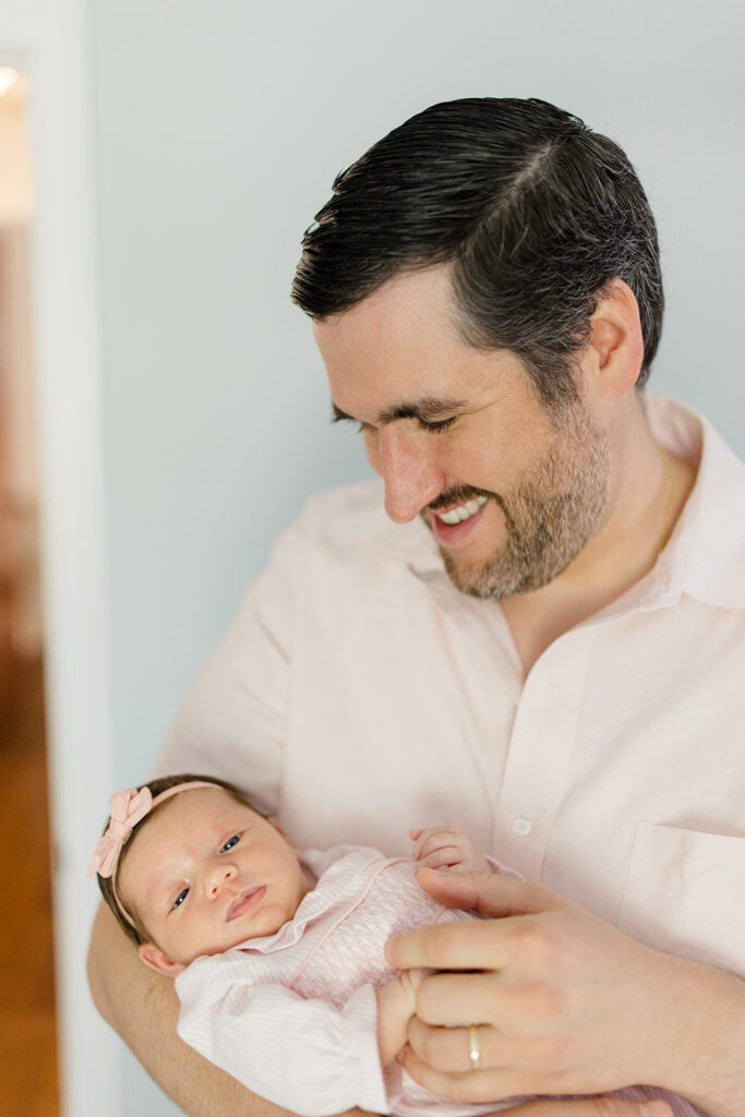Father holds his newborn in a soft pink outfit during an in-home session, wearing a light neutral shirt that provides timeless what to wear inspiration for newborn photos by a Cleveland family photographer.