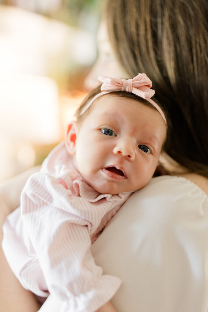 Awake newborn wearing a pink bow headband rests on her mother's shoulder while looking toward the camera in soft natural window light.