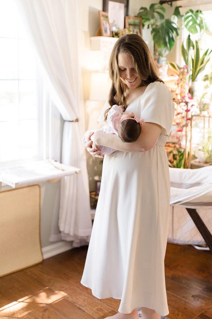 Mother smiles down at her newborn while gently rocking her in a bright, plant-filled nursery during an in-home newborn photography session.