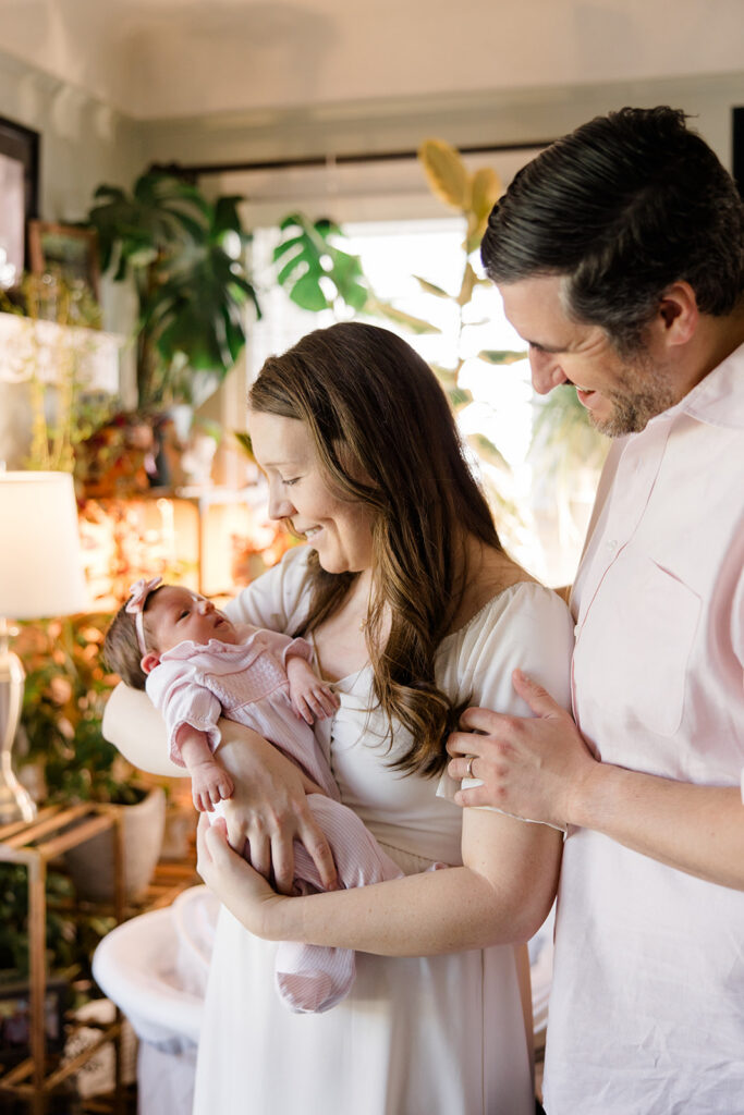 mother and father stand in their living room during newborn photos with the mother holding the baby and smiling captured by cleveland family photographer