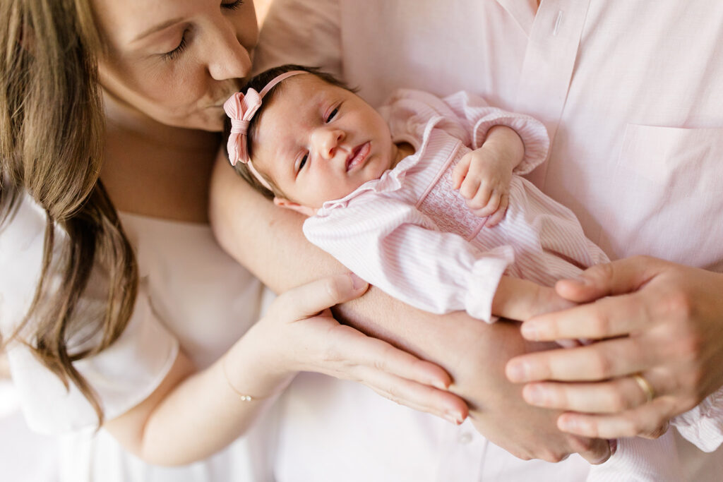Mother kisses her newborn's forehead while both parents cradle their baby together, creating a tender family moment during an in-home newborn session.