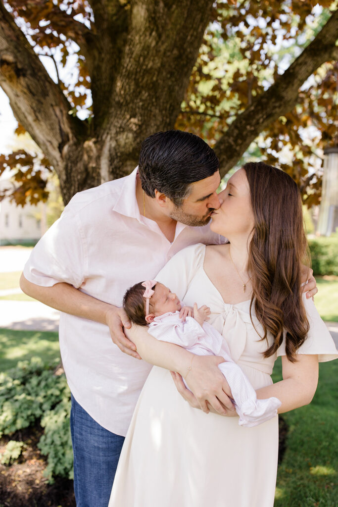 Parents share a kiss while holding their newborn outdoors, wearing coordinated neutral outfits that provide timeless what to wear inspiration for newborn photos by a Cleveland family photographer.
