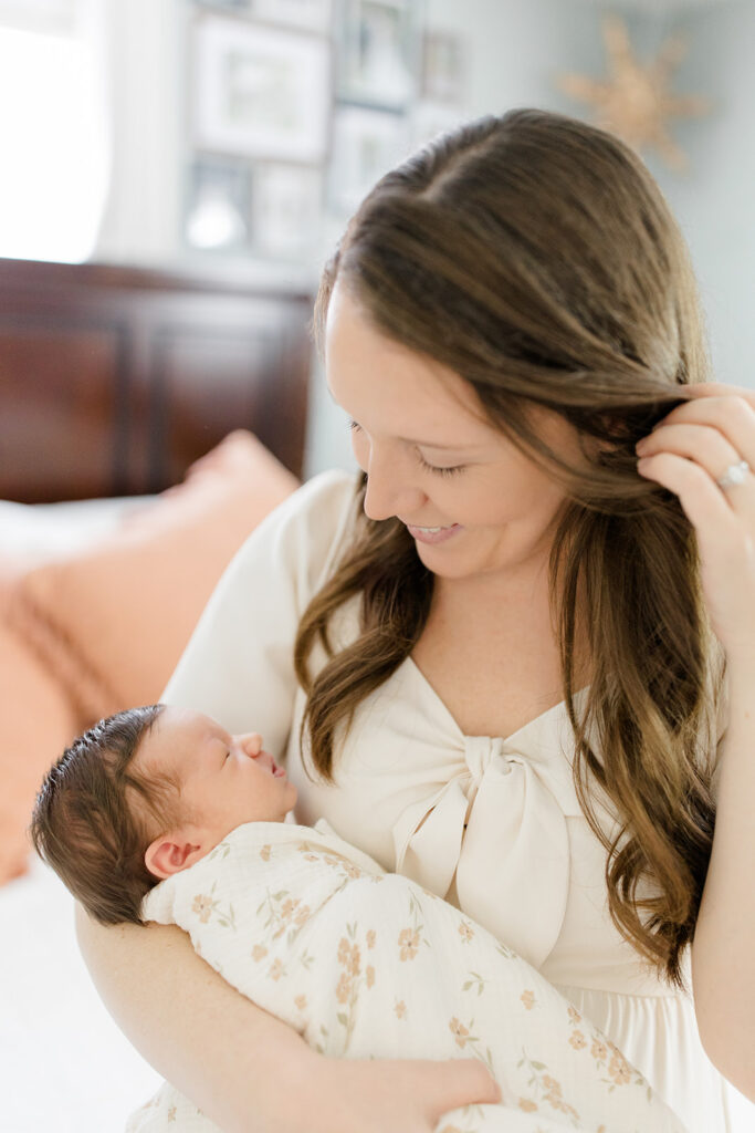 mother holding her newborn and smiling at the baby while tucking her hair behind her ear while sitting on the side of a bed