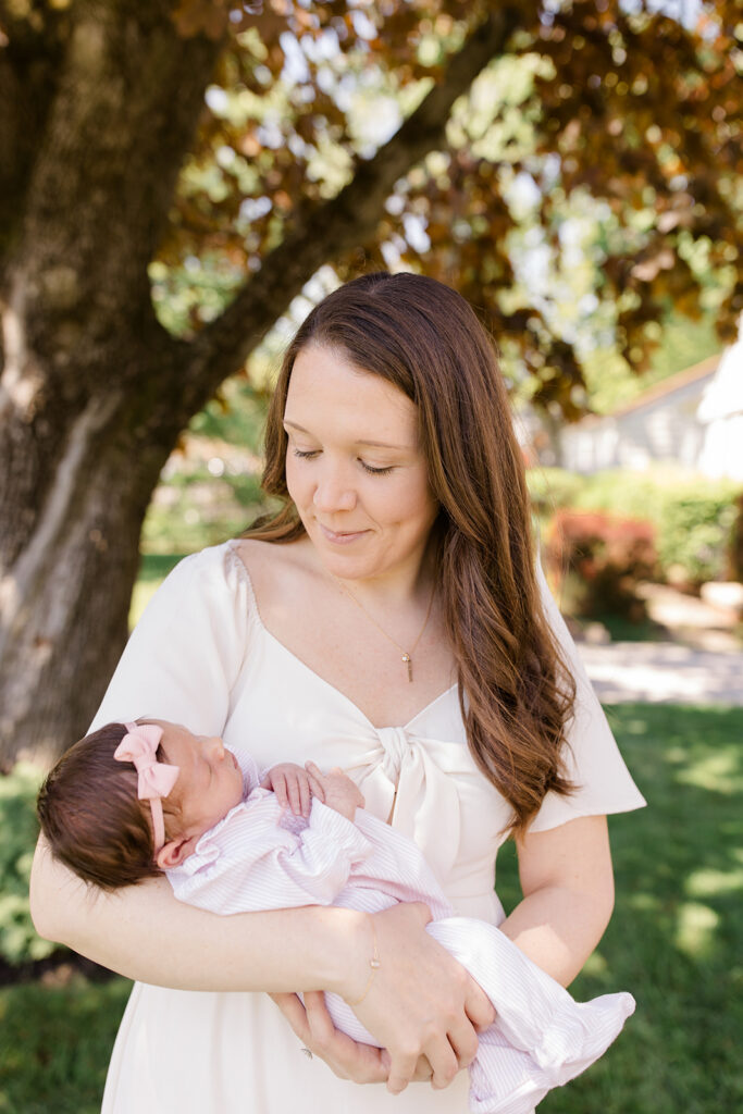 Mother cradles her newborn beneath a shady tree, wearing a flowing neutral dress that offers classic what to wear inspiration for outdoor newborn photos by a Cleveland family photographer.