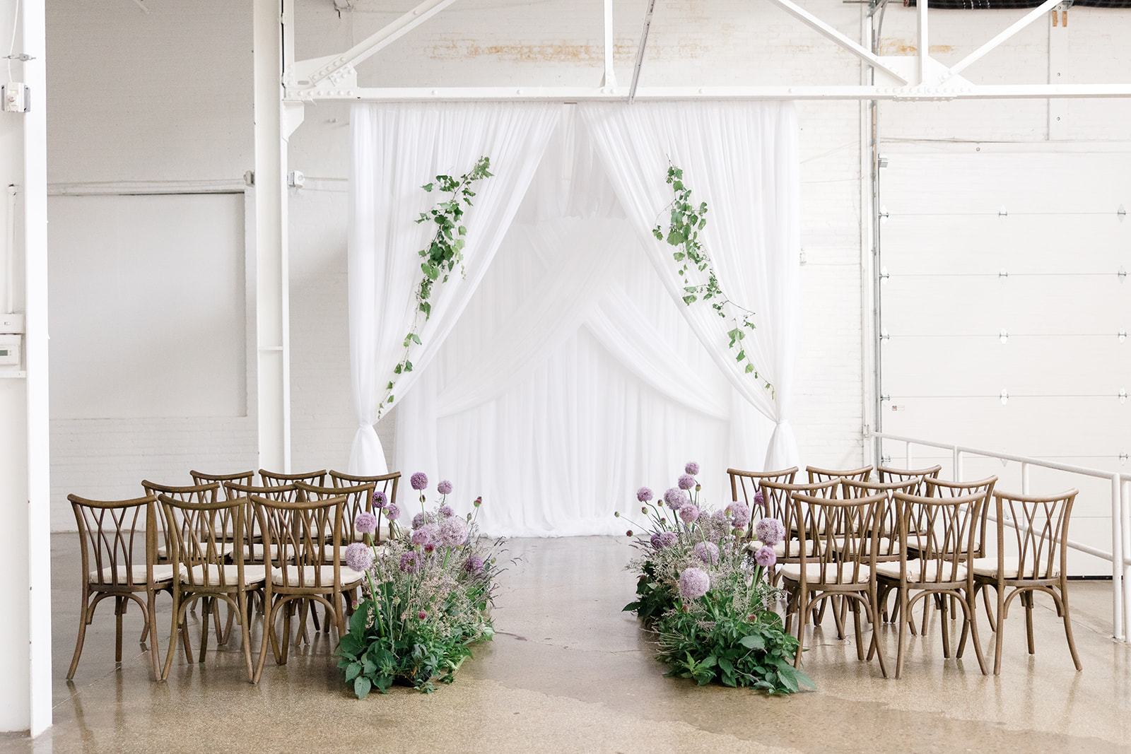 Indoor wedding ceremony setup with wooden chairs arranged in rows facing a white draped altar adorned with green vines, flanked by lush purple allium flower arrangements on both sides of the aisle, styled by a Cleveland wedding planner and photographed by a Cleveland wedding photographer.