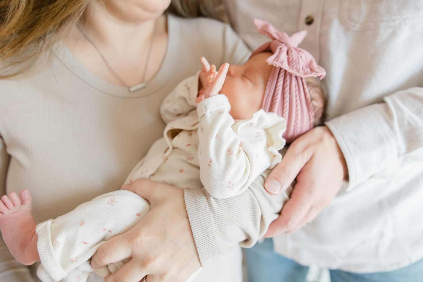 A close-up of parents holding their newborn baby dressed in a soft cream outfit with a pink headband, captured during an in home newborn photography session by a Cleveland newborn photographer.