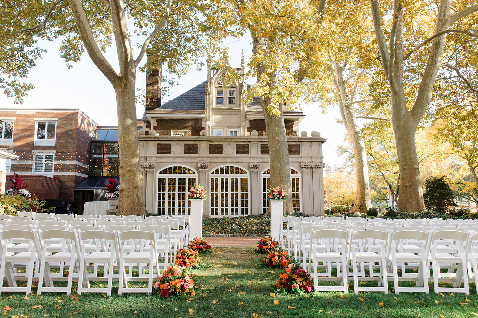 Cleveland wedding photographer captures Outdoor Glidden House wedding ceremony setup featuring rows of white chairs, floral arrangements with vibrant orange, pink, and red flowers lining the aisle, and a historic stone mansion surrounded by tall trees with autumn foliage in the background.