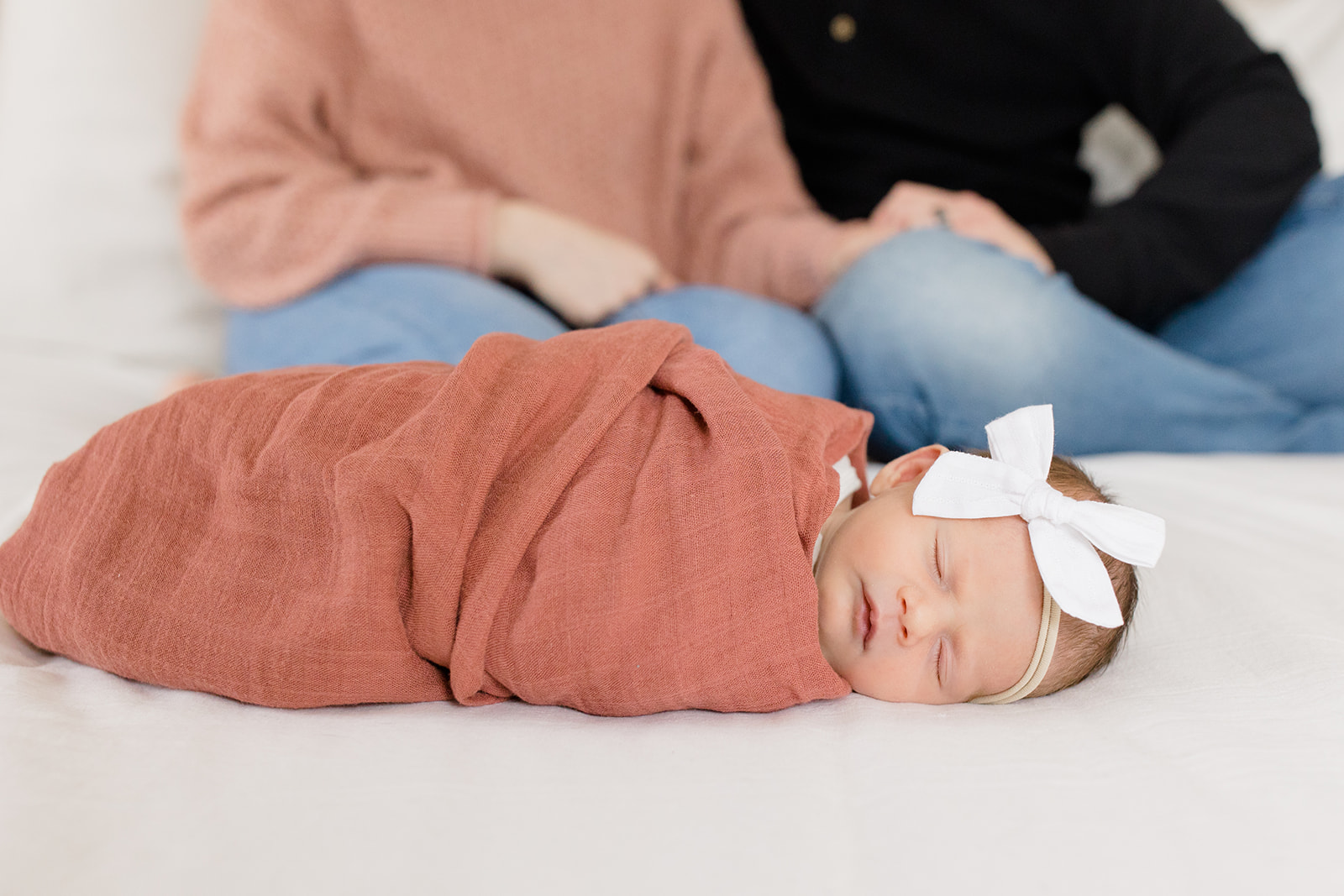 Newborn baby wrapped in a rust-colored blanket with a white bow headband, sleeping on a white bed, with parents sitting in the background during a newborn photo session by Cleveland Newborn Photographer.
