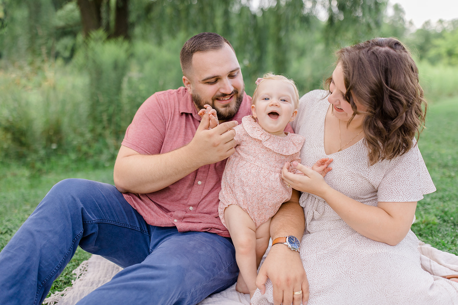 Family sitting on a blanket during a Cleveland family photography session, with the father and mother holding their smiling toddler and playing together outdoors.