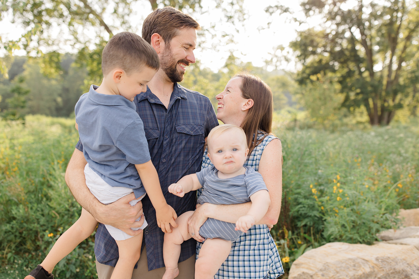 Outdoor family picture ideas with parents holding their two young children in a sunny field, smiling at each other while one child looks at the camera, captured by Cleveland family photographer.