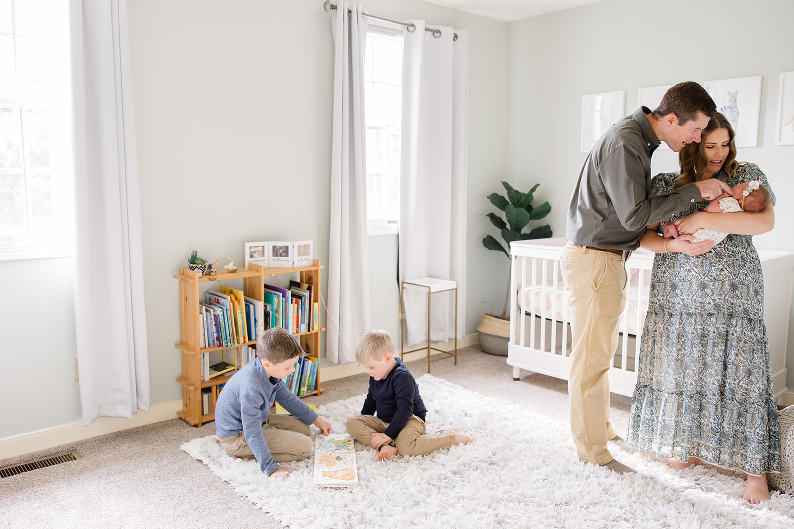 Newborn photos with siblings captured by a Cleveland family photographer showing two young boys sitting on a plush white rug reading a book, while their parents stand nearby admiring their newborn baby girl in a softly lit, neutral nursery.