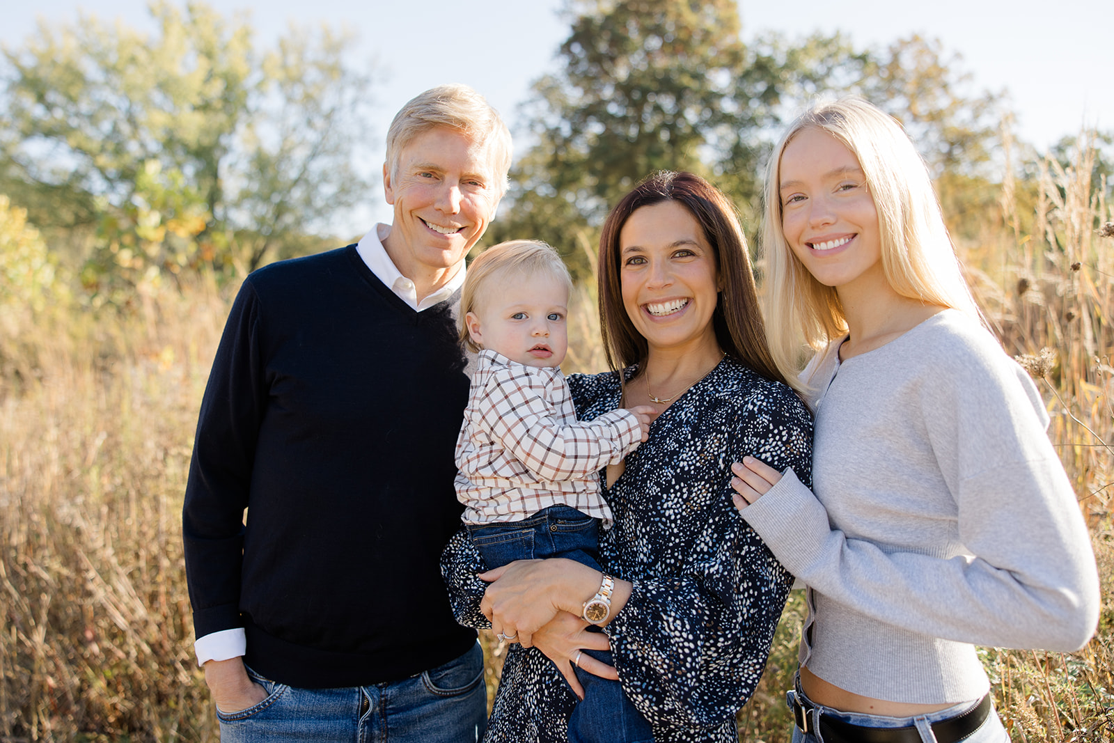 A smiling family of four poses outdoors in a sunlit field, dressed in fall attire. The mother holds a young toddler while standing beside her partner and an older daughter. Although the family dog isn’t visible in this photo, it’s part of a charming family photo session by a Cleveland family photographer.