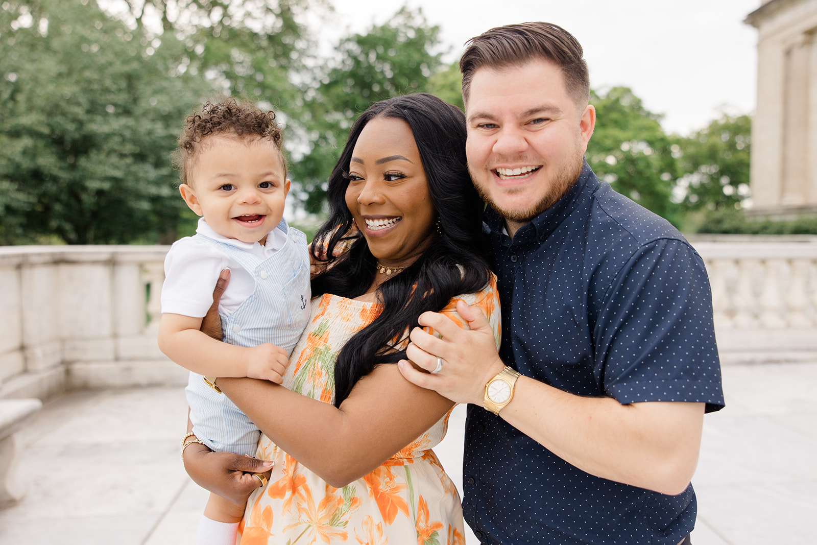 A joyful family of three poses outdoors in front of a stone terrace with trees and classical architecture in the background. The mother holds their smiling baby boy while the father hugs them both closely. This cheerful moment captures great outdoor family photo ideas by a Cleveland family photographer