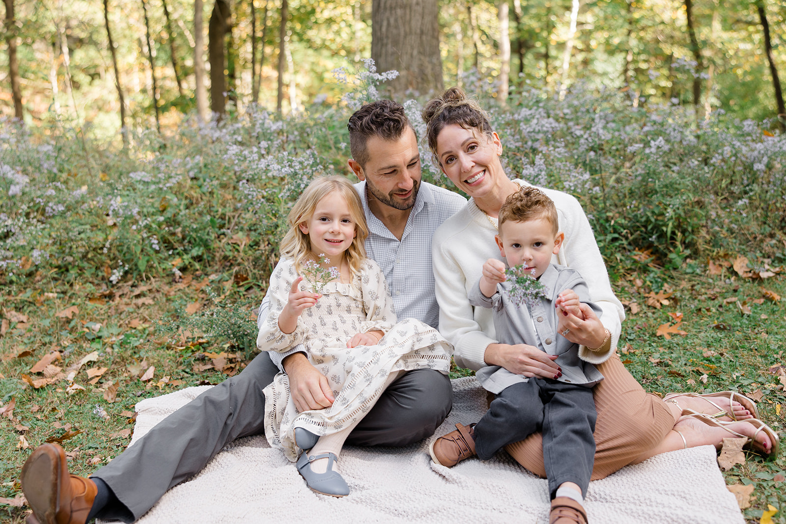 A family of four sits together on a cozy blanket in a wooded area, surrounded by wildflowers and fallen leaves. Their coordinated neutral outfits—featuring soft creams, grays, and earth tones—are perfect examples of fall family photo outfits. Captured by a Cleveland family photographer, the scene radiates warmth and connection amidst the autumn backdrop