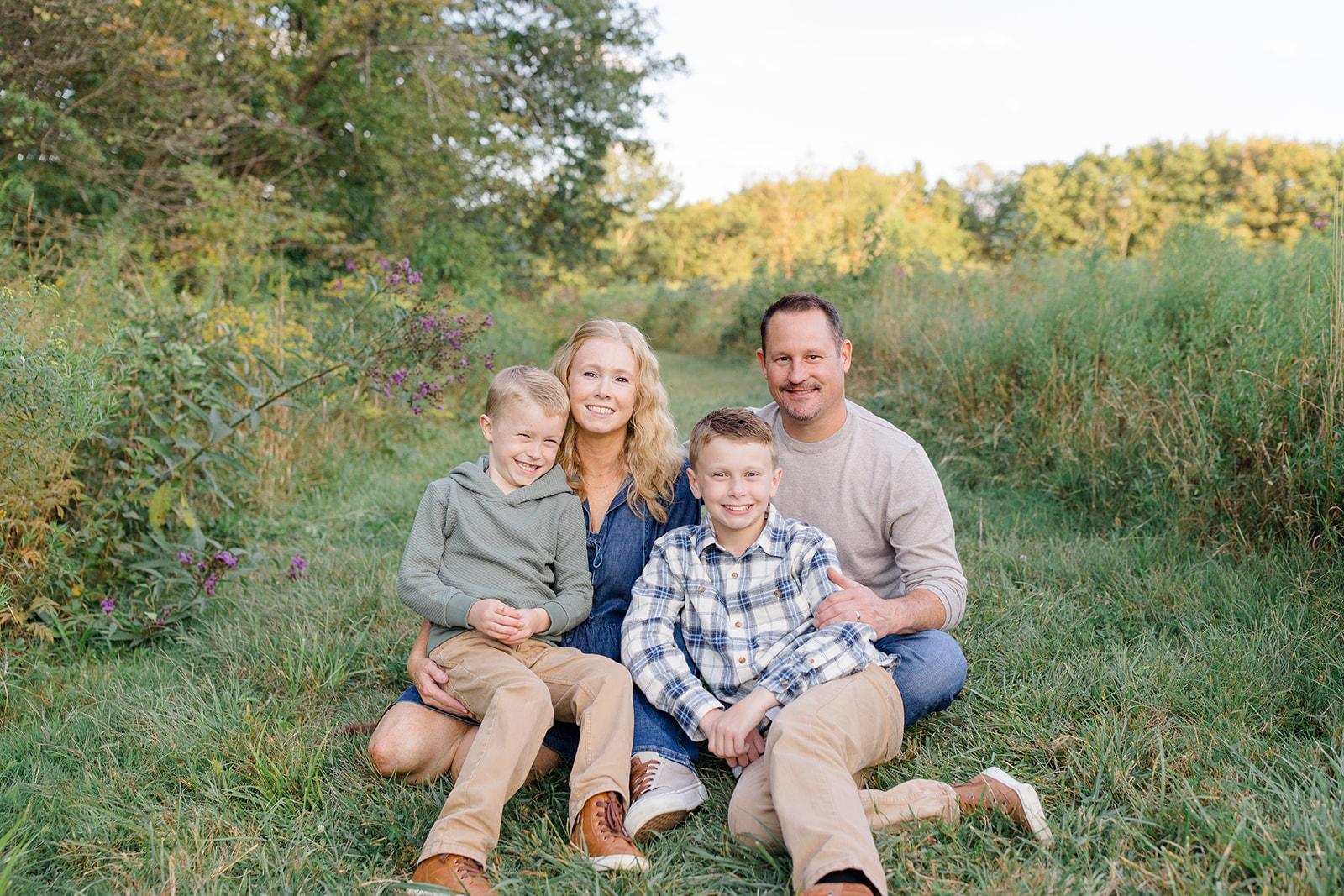 A family of four sits together in a grassy field surrounded by tall greenery and wildflowers during golden hour. The parents and two young boys smile warmly at the camera, dressed in casual fall outfits with earth tones and soft blues—perfect styling for Cleveland family photos