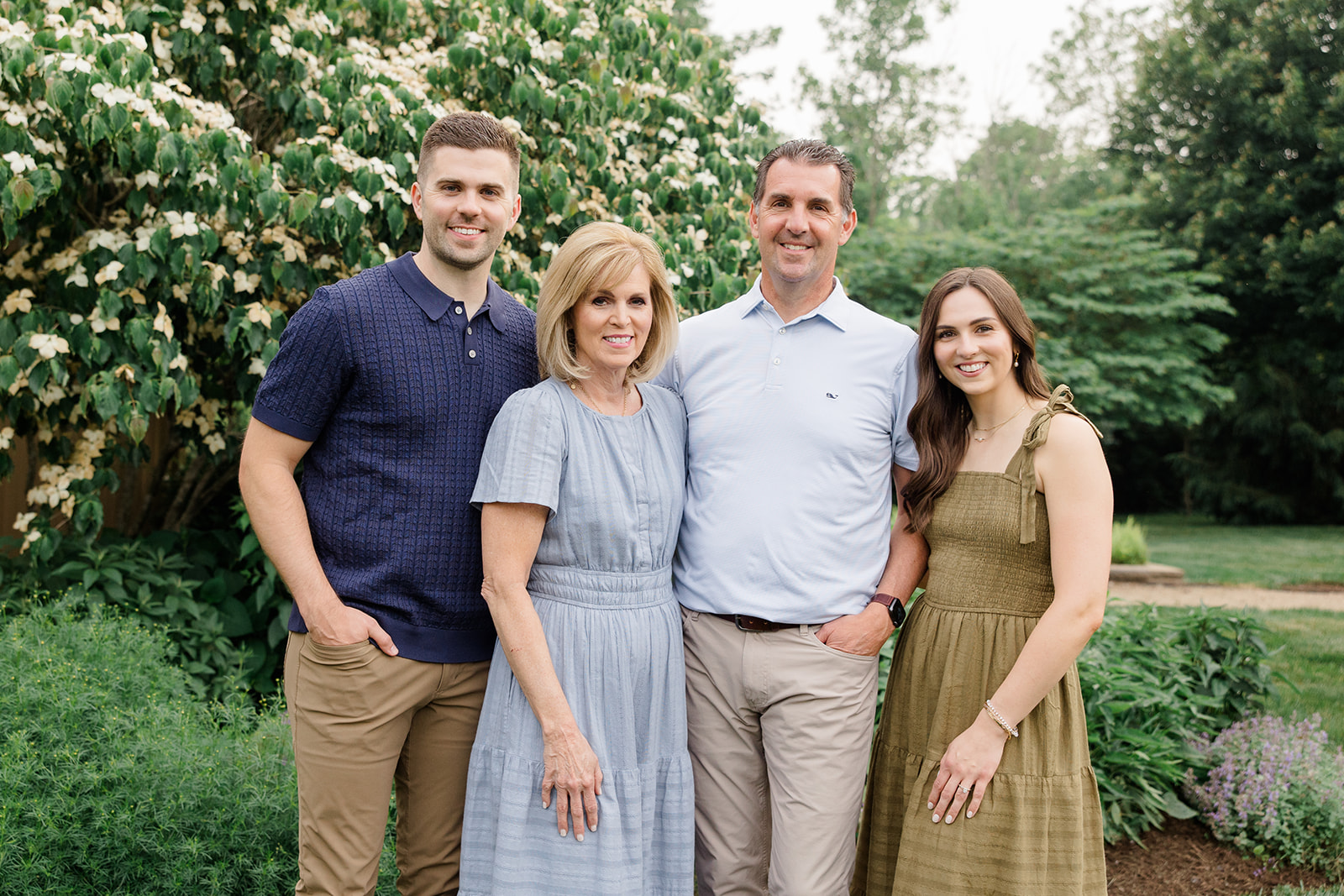 Family of four standing together in a lush garden with flowering greenery, smiling at the camera in a natural portrait showcasing Outdoor Family Photoshoot Ideas by a Cleveland family photographer.