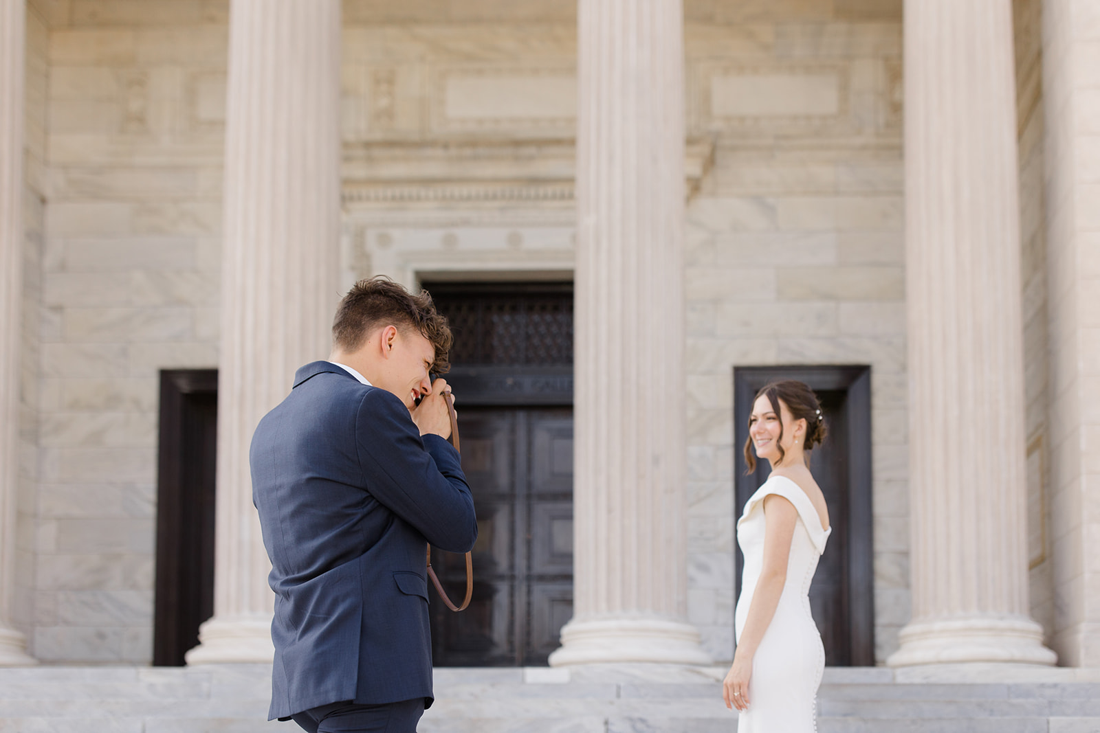 Groom photographing his bride as she smiles in front of a grand columned building, candid wedding moment captured by a Cleveland Wedding Photographer.
