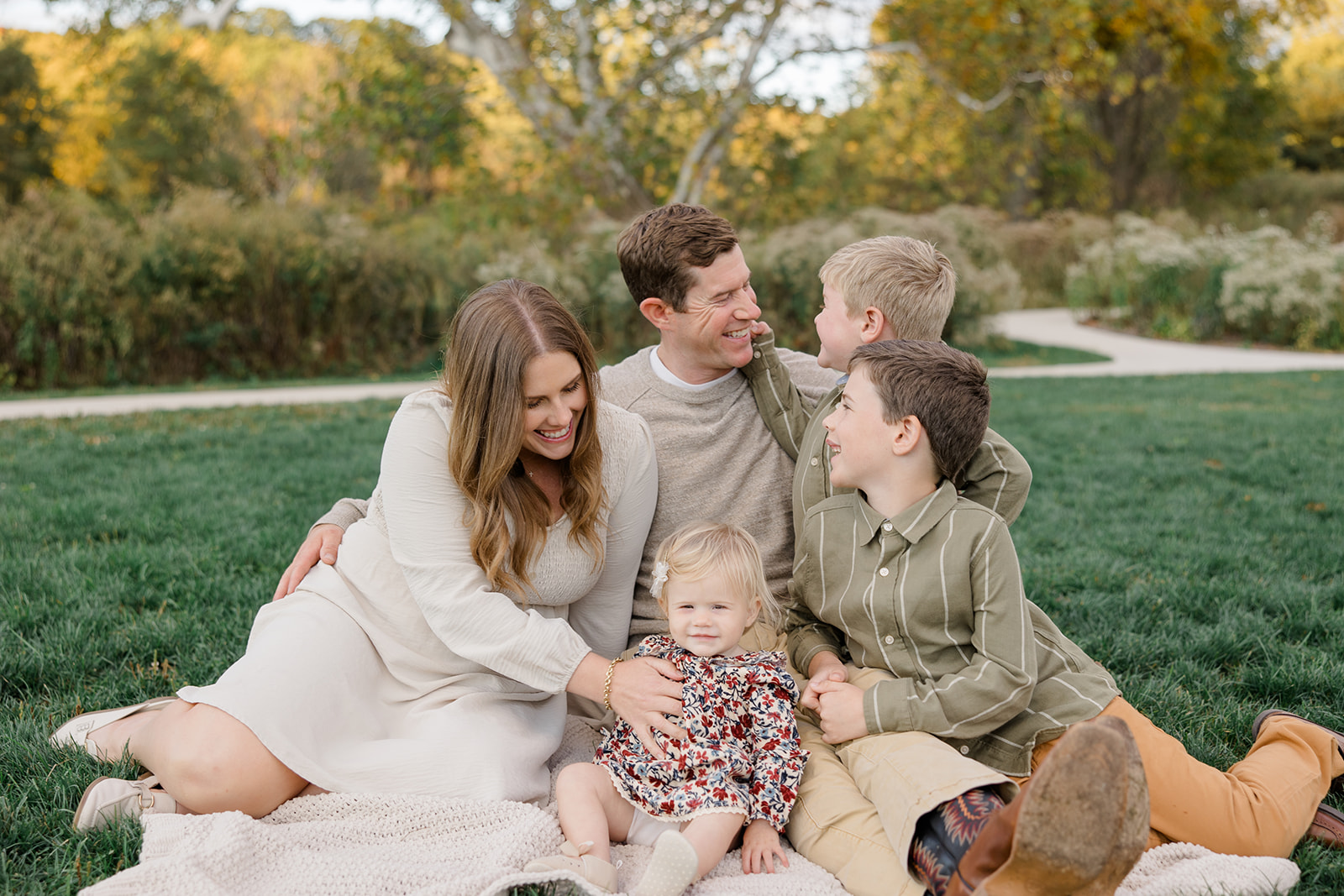 A family of five sits together on a blanket in a grassy park, laughing and interacting during an outdoor session with a Cleveland family photographer, featuring coordinated fall family picture outfits in soft neutral tones.