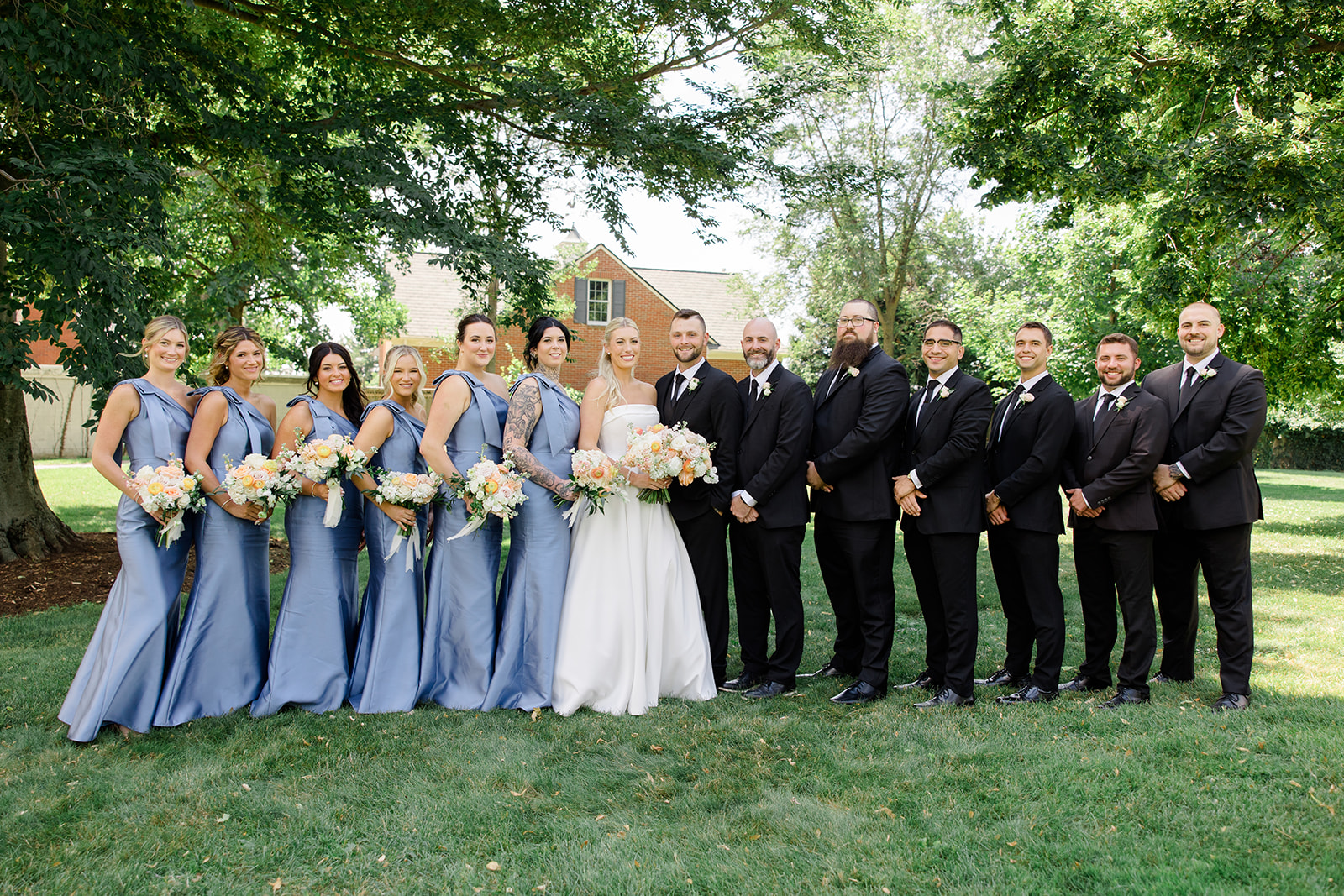 Bride and groom pose with their wedding party beneath mature trees on the lawn, with bridesmaids in blue dresses and groomsmen in black suits at one of the elegant Cleveland wedding venues. Day of Wedding Timeline